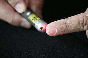 The image is a close-up shot of a person holding a small, cylindrical device that emits a bright red laser light. The device is a small, black cylinder. A bright, focused red laser beam is emanating from the end of the cylinder.  The red laser light is slightly shining through the finger at the end of the laser pointer.