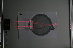 The image shows an enlarged, stylized cross-section of a human eye. On the left side, several parallel red laser beams are projected from the left and pass through a transparent block, representing the convex lens of an eye. After passing through this block the beams enter a circular area representing the eye. All beams meet at a single spot on the right side within the eye outline. 