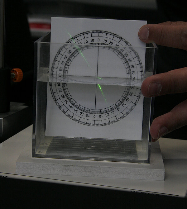 Image of a transparent water tank with a measuring scale partially submerged in the water. A beam of laser light enters the tank from the air and bends as it passes into the water, demonstrating refraction. The laser changes direction at the air-water interface, illustrating how light slows down and shifts its path when moving from air into water. The setup visually explains the principle of light refraction and the measurement of angles using the submerged scale in physics experiments
