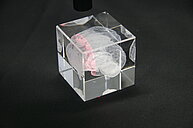 A small black laser pointer or pen is placed above a transparent acrylic cube. Inside the cube, there’s a white 3D-engraved model of a human brain. The laser beam creates a visible red line through the cube, illuminating part of the brain design. The cube is placed on a black surface.