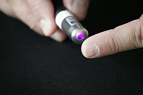 The image is a close-up shot of a person holding a small, cylindrical device that emits a bright purple laser light. The device is a small, black cylinder with a white band near the top. A bright, focused purple laser beam is emanating from the end of the cylinder.   
