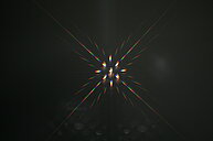 The image depicts a dazzling burst of light against a completely black background. It appears to be a photograph taken looking directly at a very bright light source, possibly a street lamp or a bright bulb. The light source is located in the center of the frame, and its rays radiate outwards in all directions, creating a starburst effect. The rays are multicolored, displaying a spectrum of colors including red, orange, yellow, green, blue, and purple, although the dominant colors seem to be red, green, and blue. The intensity of the light is so high that it creates lens flares and diffraction spikes, adding to the complexity and brilliance of the image. The overall impression is one of intense brightness and vibrant color against a stark, dark void.
