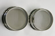 The image shows two identical metal strainers or sieves placed side-by-side on a white surface. Each strainer is round with a slightly raised rim and a handle extending upwards from the side. The mesh inside the strainers is fine and made of thin, silvery wire, creating a pattern of small, evenly spaced holes. The metal of the frame and handle appears to be stainless steel or a similar shiny metal. The background is a plain white surface, providing high contrast with the metal objects. The overall composition is simple, focusing on the texture and form of the strainers.