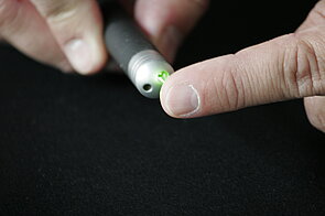 The image is a close-up shot of a person holding a small, cylindrical device that emits a bright green laser light. The device is a small, black cylinder. A bright, focused green laser beam is emanating from the end of the cylinder.   