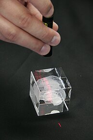 The image shows a person holding a small black laser pointer or pen above a transparent acrylic cube. Inside the cube, there’s a white 3D-engraved model of a human brain. The laser beam creates a visible red line through the cube, illuminating part of the brain design. The cube is placed on a black surface.