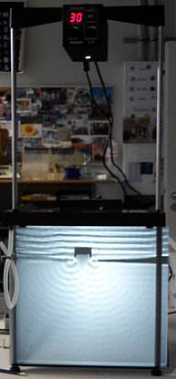 Image of a ripple tank, a shallow transparent tray filled with water used to study wave behavior. The tank shows waves generated on the water surface, illustrating phenomena such as reflection, refraction, diffraction, and interference. Light shining through or above the water enhances the visibility of wave patterns.