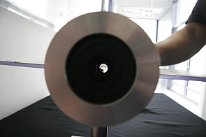 The image is dominated by a large metallic disc positioned centrally in the foreground, seen head-on so that its perfectly round shape fills much of the frame. In the middle of the disc is a black circular opening, which appears to be the lens or aperture of an optical device. Through this central opening, some light is visible, creating small reflections. To the right side of the image, a person’s arm is extended, partly visible and slightly out of focus against a bright background. The scene is set indoors, with windows and daylight visible in the background, and a dark cloth or surface beneath the disc.