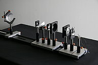 The image displays an optical setup featuring six distinct components arranged linearly on a black surface. Each component is mounted on an adjustable stand with an orange knob. From left to right, the components include a large lens/mirror assembly, a square dichroic mirror/filter, a smaller convex lens, another square lens/filter, and finally a light source module.