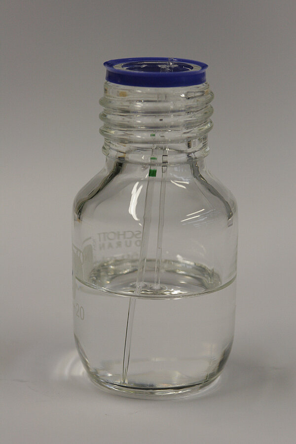 Image illustrating light refraction with a half-filled bottle of water. Two pipettes stand upright in the bottle: one is filled with water and becomes nearly invisible underwater due to the matching refractive index, while the other pipette is filled with air and remains clearly visible.